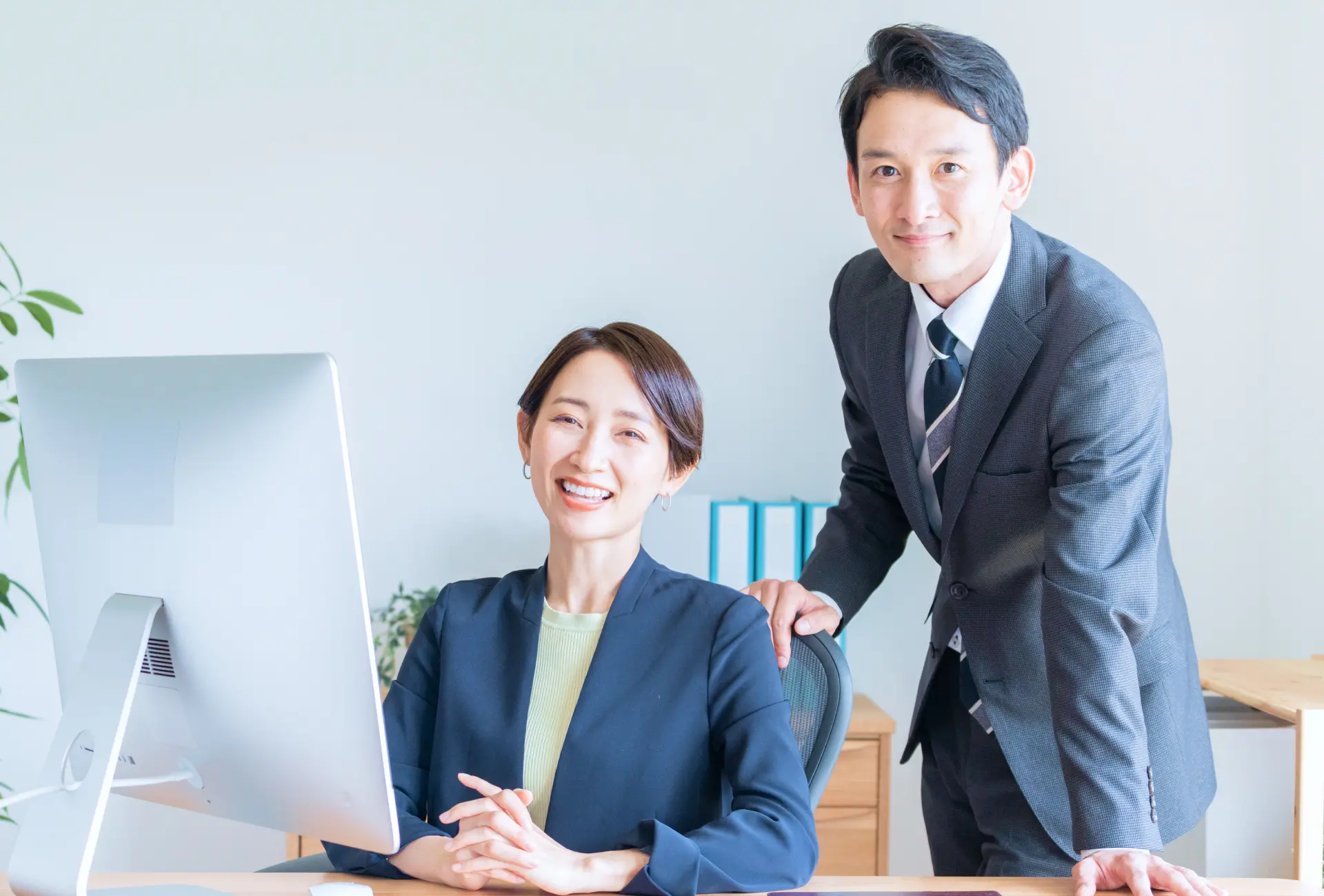 パソコンが置かれたデスクの後ろに座っている女性社員と椅子に手をついている男性社員の写真